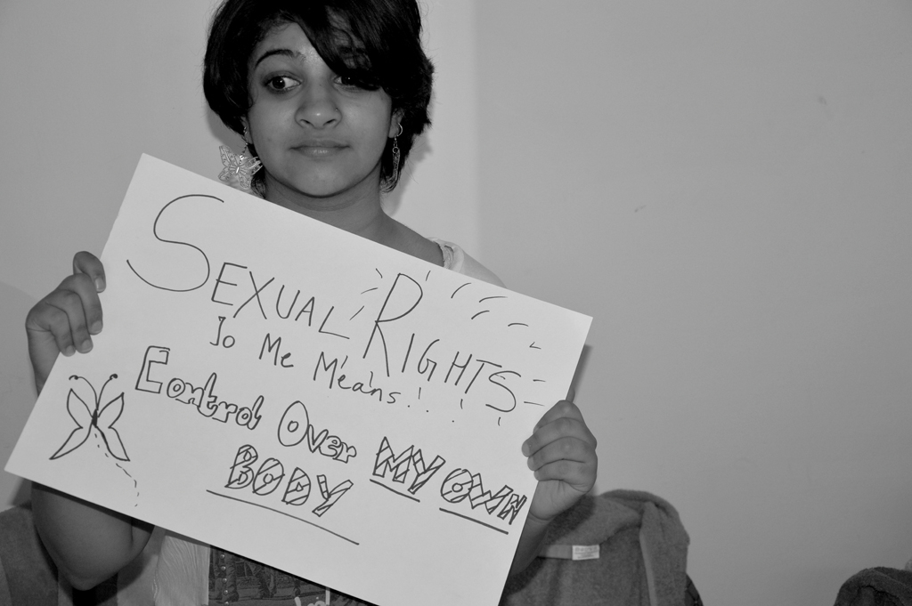 A person with short dark hair and silver earrings holds a sign that says "Sexual Rights To Me Means Control Over My Own Body". The photo is in black and white.