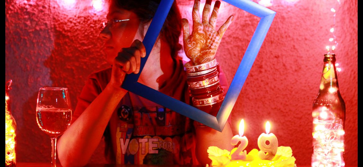 A person with dark hair and glasses holds up a blue picture frame to display the mehendi on their hand and the chura on their wrist. A birthday cake with the number 29 is to the right, and a wine glass is to the left. The person's head is turned to the side; the backdrop is a red wall.
