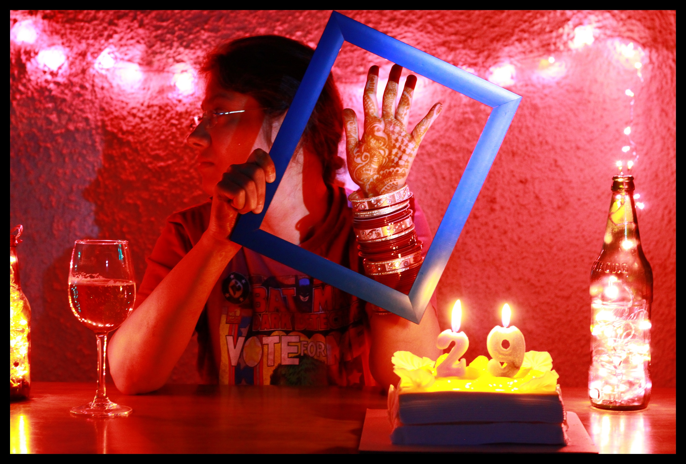 A person with dark hair and glasses holds up a blue picture frame to display the mehendi on their hand and the chura on their wrist. A birthday cake with the number 29 is to the right, and a wine glass is to the left. The person's head is turned to the side; the backdrop is a red wall.
