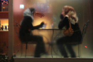 Two people wearing hijabs sit at a small table behind a foggy window