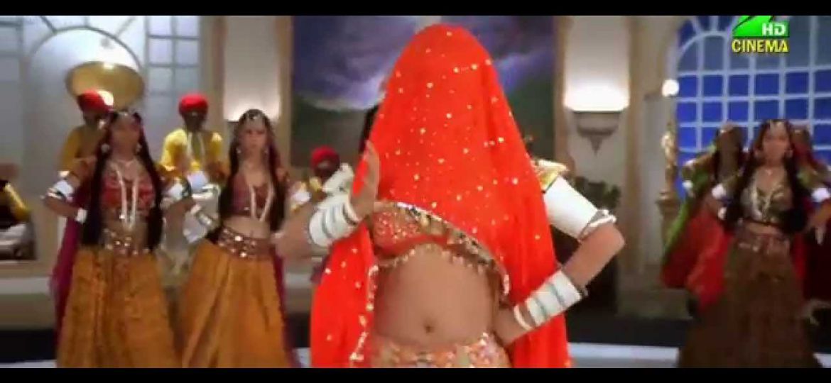 A still from the song "Choli Ke Peeche Kya Hai" - a person wearing white bangles and an orange lehenga is dancing, with background dancers wearing brown lehengas in the back. An orange dupatta covers the person's head.