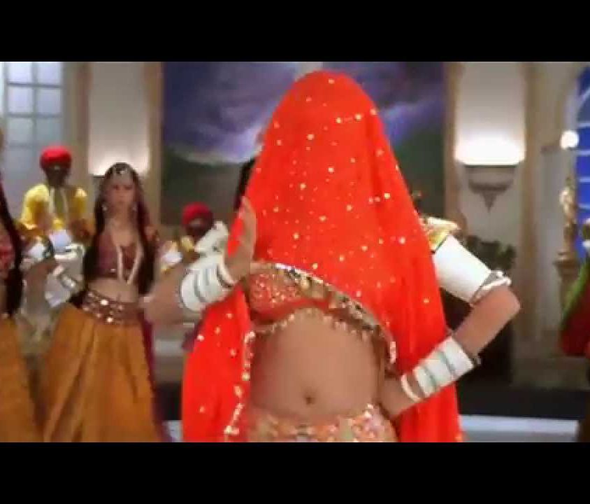 A still from the song "Choli Ke Peeche Kya Hai" - a person wearing white bangles and an orange lehenga is dancing, with background dancers wearing brown lehengas in the back. An orange dupatta covers the person's head.