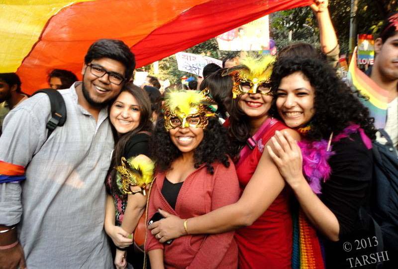 Five smiling people posing in front of a waving rainbow flag. Two are wearing gold masks with yellow feathers. Source: TARSHI