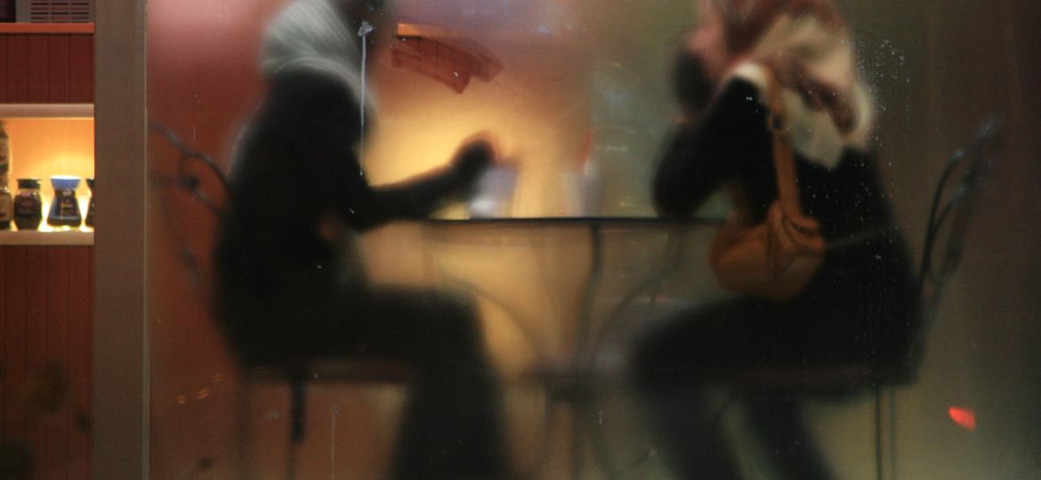 Two women wearing full black clothing and a headscarf sitting in a cafe. The photo is taken from other side of a glass door, such that they are blurred.