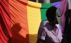 A photo of a person in a black mask and dressed in a purple shirt, standing in front of a rainbow-coloured pride flag.