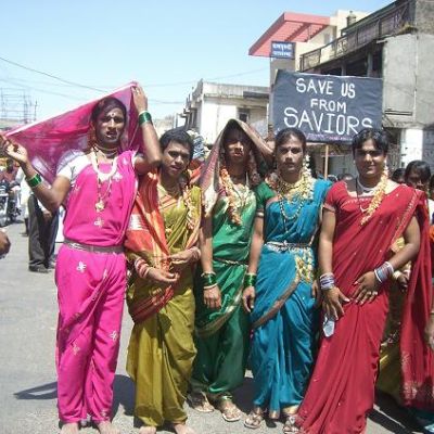 Five transgender people walking down on an Indian road. Someone behind them is carrying a placard "Save us from saviors". They are wearing bright-coloured sareers, gajras, bindis, and jewelery.