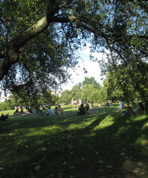 A park in which multiple couples are seen sitting together at various points. There are two big trees on each side, casting shadows on the same-sex couple sitting in the very centre with their backs turned.