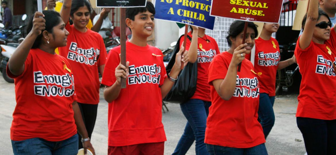 Youngsters wearing red shirts with "Enough is enough" written in black march on a road, carrying placards reading "Child sexual abuse - what are you doing about it?"; "The most public secret needs to be told"; and "it takes a community to protect a child."