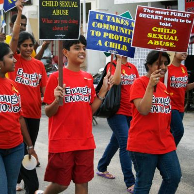 Youngsters wearing red shirts with "Enough is enough" written in black march on a road, carrying placards reading "Child sexual abuse - what are you doing about it?"; "The most public secret needs to be told"; and "it takes a community to protect a child."
