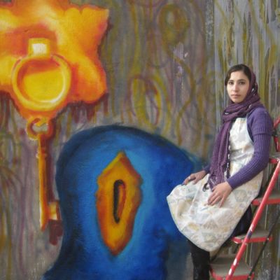 Picture of Afghani street artist Malina Suliman sitting in front of one of the street murals she has painted. She is wearing a white kurta with a purple cardigan and a purple headscarf.