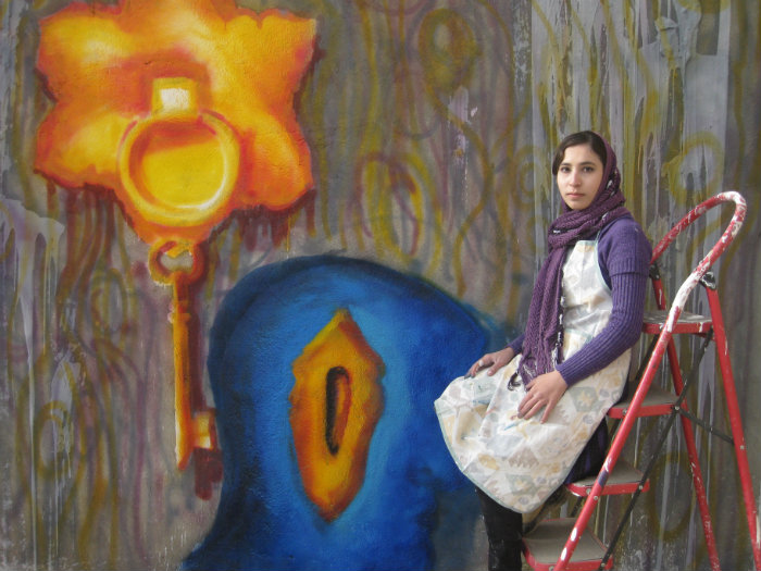 Picture of Afghani street artist Malina Suliman sitting in front of one of the street murals she has painted. She is wearing a white kurta with a purple cardigan and a purple headscarf.