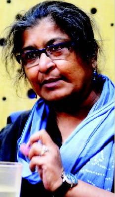 A picture of feminist scientist Chayanika Shah. Her hair is dark, with specks of grey. She has a blue scarf wrapped around her neck, and is wearing blue earrings.