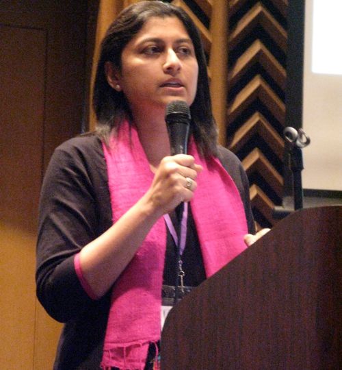 Photo of safe abortion advocate Suchitra Dalvie. She is standing near a podium, holding up a mic. She is wearing a black kurta and a pink scarf around her neck.