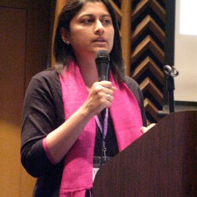 Photo of safe abortion advocate Suchitra Dalvie. She is standing near a podium, holding up a mic. She is wearing a black kurta and a pink scarf around her neck.