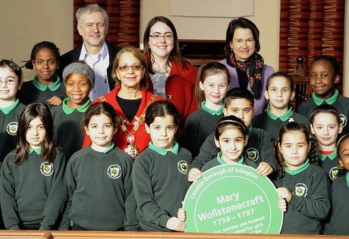 A picture of a class full of young students with their four teachers standing at the back. One of the students towards the front is holding up a placard with Mary Wollstonecraft's name written on it.