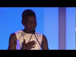 Photo of actress Lupita Nyongo speaking from a podium. She is wearing a white dress.