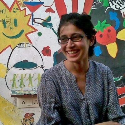 Photo of bodypositive activist Ekta Oza. She is wearing dark-rimmed glasses, a blue patterned kurta and her hair is tied back. She is looking to her left and laughing.