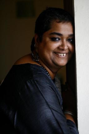 Photo of queer feminist activist Pramada Menon. She has short hair and is leaning against a wall. She is wearing a blue kurta, and a necklace.