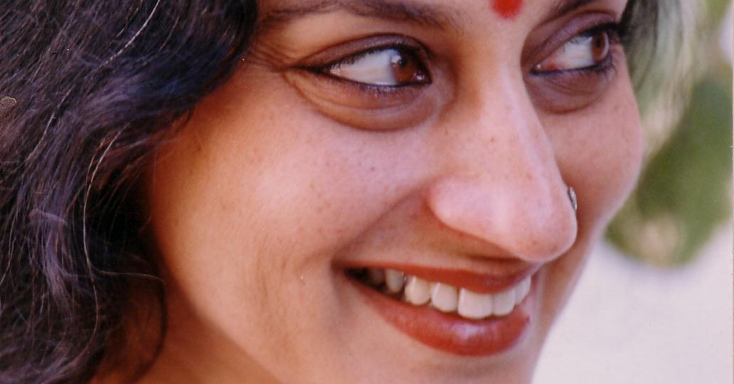 Photo of performance artist and theatre personality Maya Krishna Rao. She is wearing black and her left hand raised in a dramatic pose. She has long white hair and there is a red bindi on her forehead.