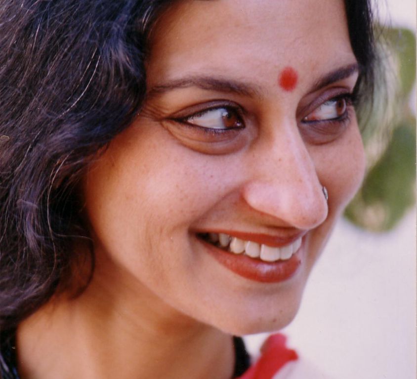Photo of performance artist and theatre personality Maya Krishna Rao. She is wearing black and her left hand raised in a dramatic pose. She has long white hair and there is a red bindi on her forehead.