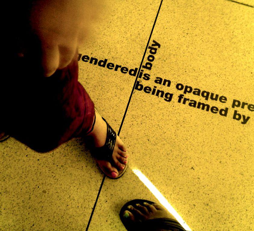 Photo shows a tiled floor lit by yellow lighting. Two different sets of feet stand facing each other.