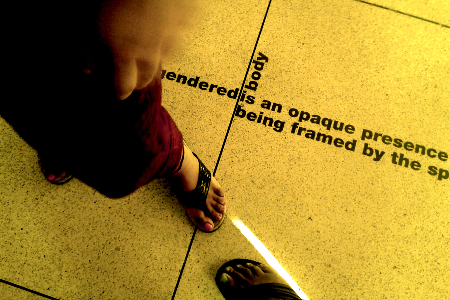 Photo shows a tiled floor lit by yellow lighting. Two different sets of feet stand facing each other.