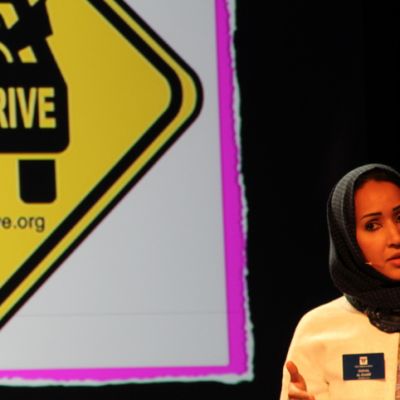 Photo of Saudi Arabian women's rights activist Manal Al Shairf. She has black hair and is wearing a black headscarf, and is standing in front of a board that says 'women2drive'.