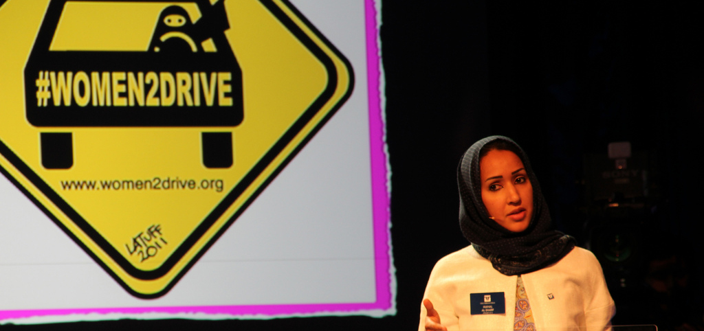 Photo of Saudi Arabian women's rights activist Manal Al Shairf. She has black hair and is wearing a black headscarf, and is standing in front of a board that says 'women2drive'.