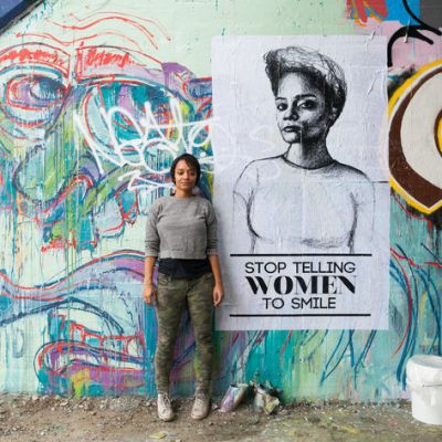 Artist Tatyana Fazlalizadeh stands infront of a wall that has a mural painted on it. She has short hair and is wearing a simple top with trousers. There is also a poster behind her that says "stop telling women to smile"