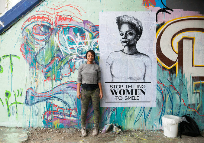 Artist Tatyana Fazlalizadeh stands infront of a wall that has a mural painted on it. She has short hair and is wearing a simple top with trousers. There is also a poster behind her that says "stop telling women to smile"