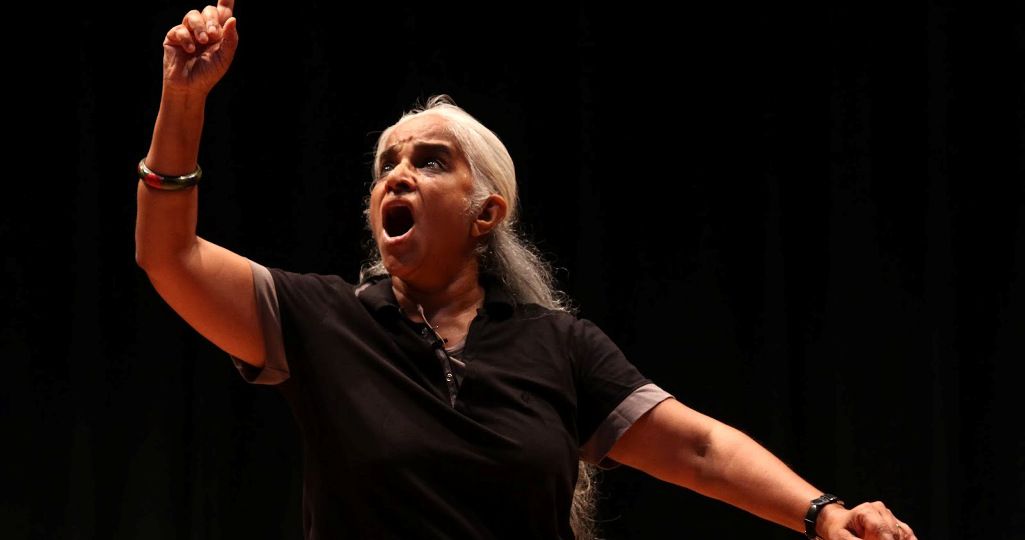 Photo of performance artist and theatre personality Maya Krishna Rao. She is wearing black and her left hand raised in a dramatic pose. She has long white hair and there is a red bindi on her forehead.