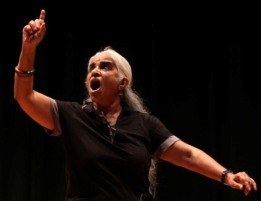 Photo of performance artist and theatre personality Maya Krishna Rao. She is wearing black and her left hand raised in a dramatic pose. She has long white hair and there is a red bindi on her forehead.