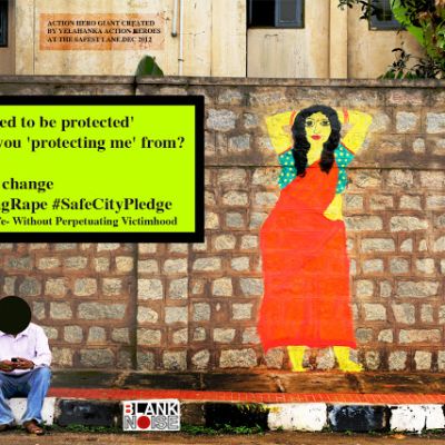 photo shows a brown wall which has an illustration of a woman dressed in a red saree and blur blouse. a man sits in fron of the wall, but his face is blacked out. The message written on the wall says, "i don't 'need to be protected'/ What are you 'protecting me' from? / Think / Make That Change #DelhiGangRape #SafeCityPledge