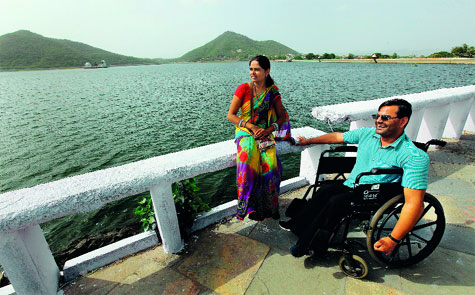 A man dressed in a light blue shirt and black trousers sits on a wheelchair. He is in a public place - a sort of a bridge, that overlooks a river. A woman dressed in a multi coloured sari sits on the railing of the bridge.