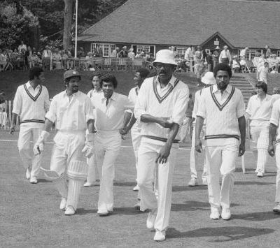 A black and white picture of an all-male cricket team. They are dressed in white jerseys with green borders and white trousers. A few of them wear caps.