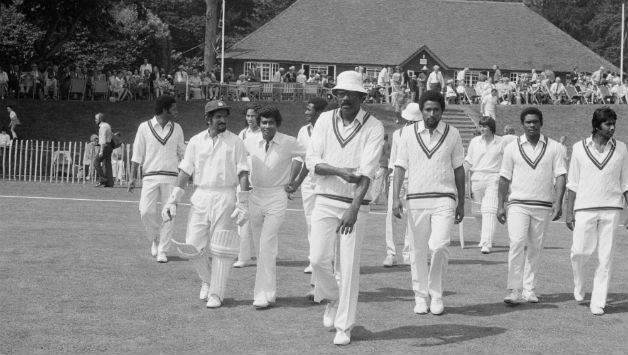 A black and white picture of an all-male cricket team. They are dressed in white jerseys with green borders and white trousers. A few of them wear caps.