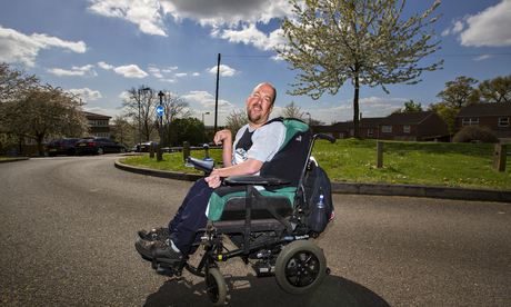 A man sits on a wheelchair. He is balding, and is wearing a white shirt and plain blue trousers.