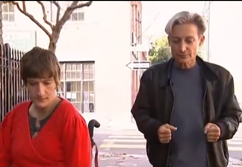 feminist theorist judith butler, wearing a black shirt and trousers, walks beside wheelchair-bound disability rights activit sunnaura taylor, who's wearing a bright red top.