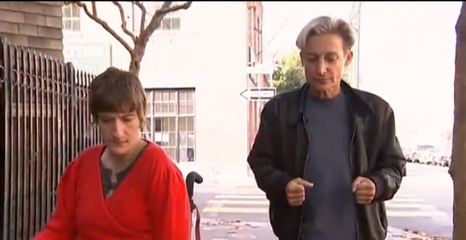 feminist theorist judith butler, wearing a black shirt and trousers, walks beside wheelchair-bound disability rights activit sunnaura taylor, who's wearing a bright red top.