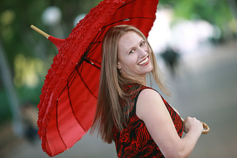 A woman dressed in a black and red dress and holding up a big red umbrella is looking behind. She has long blonde hair and a smile on her face.