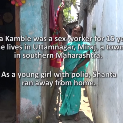 Photo of a woman in a light blue salary standing leaning against a wall in what looks like arun-down neighbourhood. The text over the image reads: "Shanta Kamble was a sex worker for 15 years. She lives in Uttamnagar, Miraj, a town in southern Maharashtra. As a young girl with polio, Shanta ran away from home."