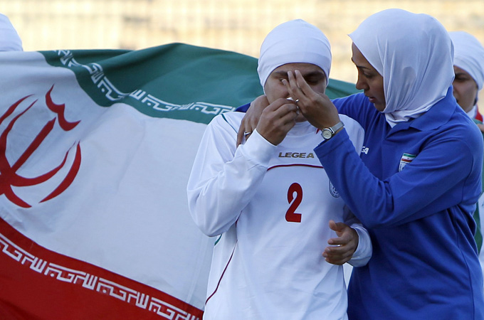 A female athlete with a headcarf around her head has her eyes closed and seems to be crying. She is being comforted by another woman in a white hijabn and a blue jersey.