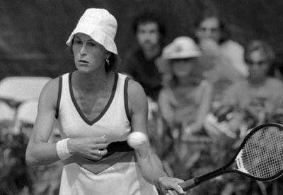 A photo of Renee Richards, the first transsexual woman to play in competitive sport after transitioning. She is wearing a white uniform and cap and is holding up a tennis racket.