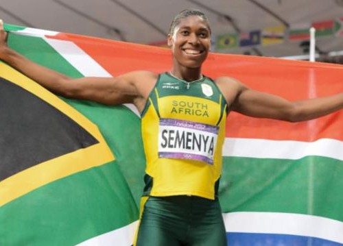 A picture of athlete Caster Semenya. She is wearing a yellow and green jersey with 'south africa' written on it and is holding up a flag of south africa behind her.