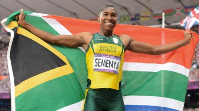 A picture of athlete Caster Semenya. She is wearing a yellow and green jersey with 'south africa' written on it and is holding up a flag of south africa behind her.