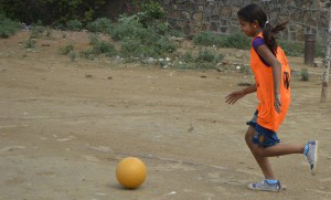 Picture of a girl playing football. She's wearing an orange jersey with purple sleeves and blue shorts. Her hair is tied back in a ponytail.