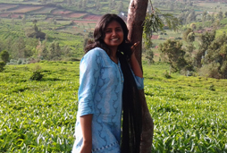 A photo of a woman standing in the middle of a green meadow, leaning against a brown tree bark. She has shoulder length black hair and is wearing a light blue kurta