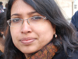 photo of activist Dr. Payoshini Mitra. She wears glasses and long hair, and is wearing a black sweater with an orange-and-black patterned scarf