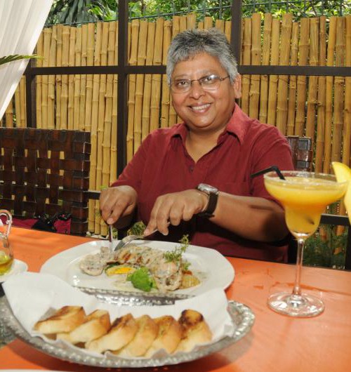 Picture of film scholar Shohini Ghosh. She's dressed in a red shirt, has white hair, and is sitting in front of a table full of food.