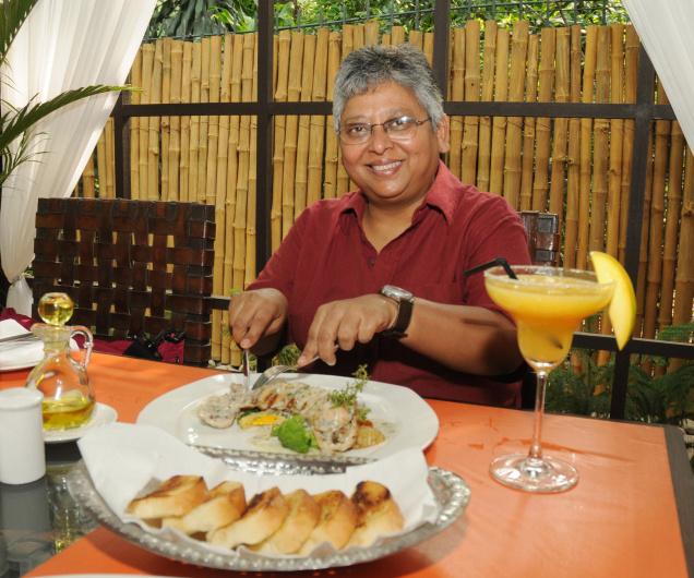 Picture of film scholar Shohini Ghosh. She's dressed in a red shirt, has white hair, and is sitting in front of a table full of food.